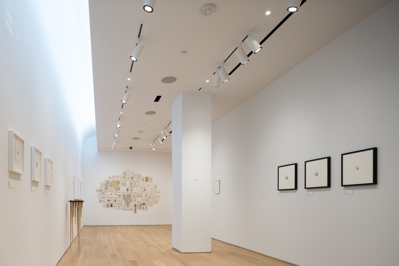 Gallery corridor with white walls, track lighting, and oak floors featuring framed artwork and central collage installation.