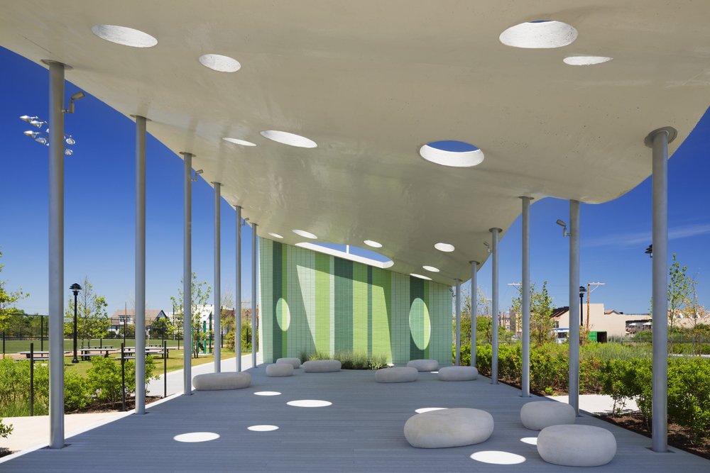 A pavilion with a perforated canopy, green glazed brick wall, and stone seating in a park.