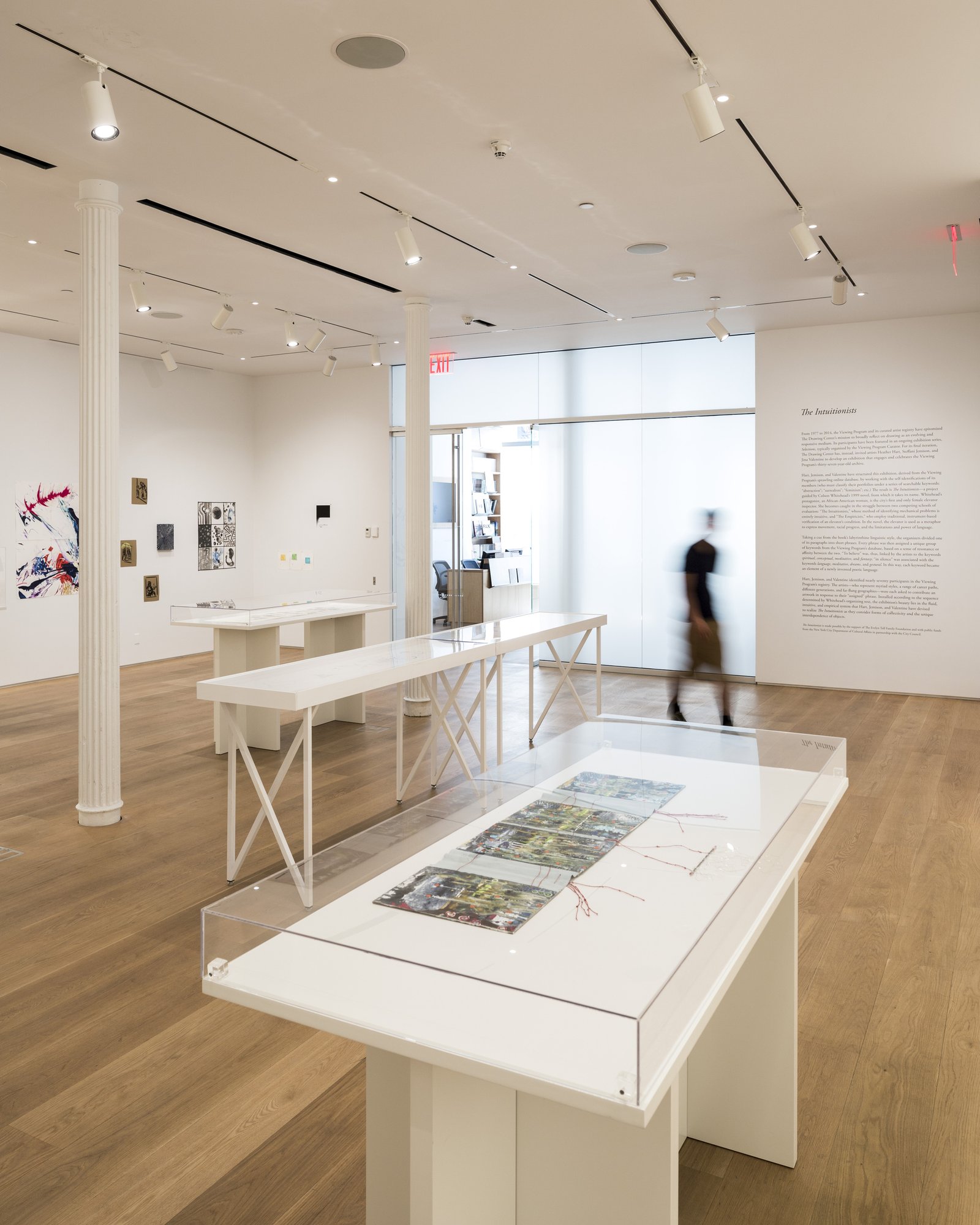 Gallery interior with white display cases, fluted column, oak flooring, and track lighting; blurred figure passing through.