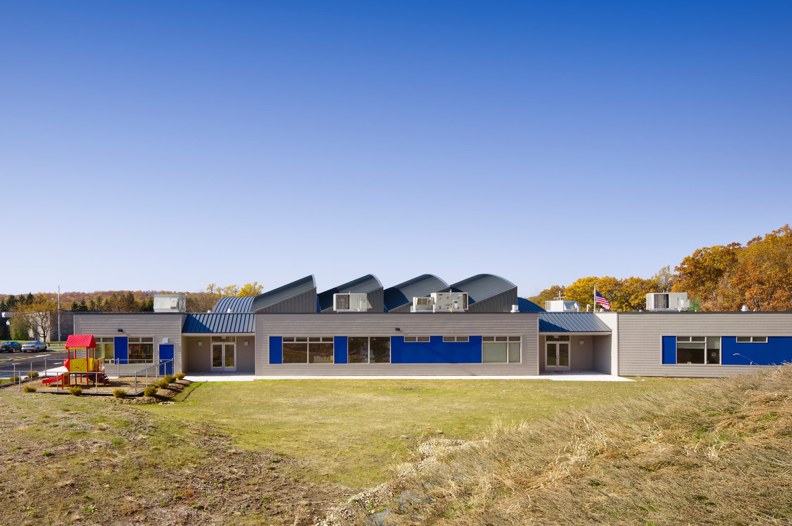 A school building with blue accents, curved roofs, and a playground, set against autumn trees.