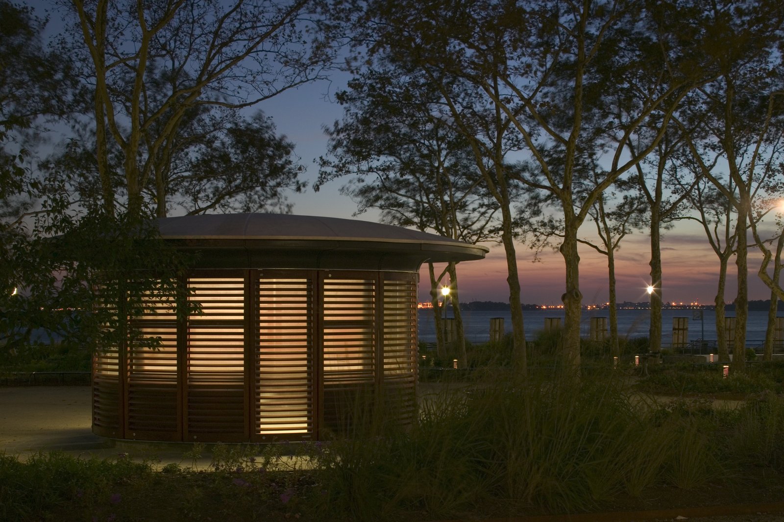 A softly lit kiosk stands among trees at dusk with the New York Harbor in the background.