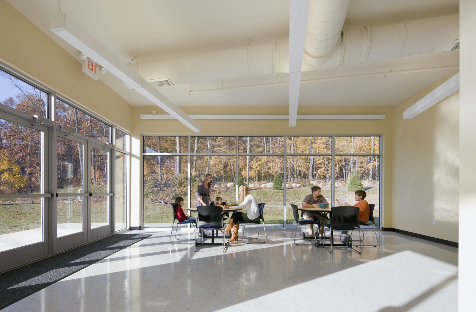 A sunlit classroom with floor-to-ceiling windows, students, and teachers engaged in activities.