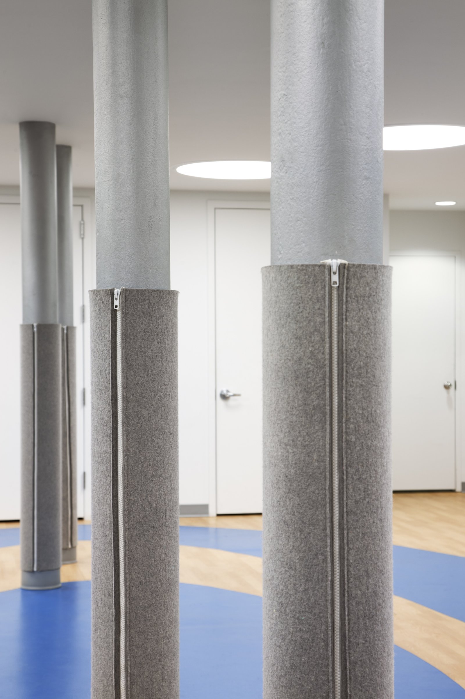 Close-up of structural columns wrapped in gray padded fabric with zippers in a blue and wood-floored space.