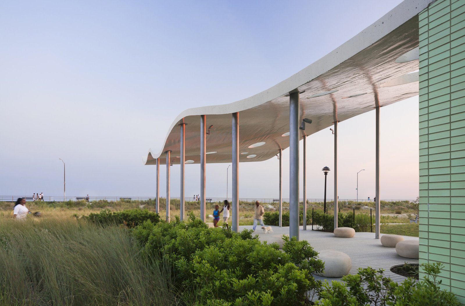 A curved pavilion canopy with perforations, adjacent to green brick walls and surrounded by coastal grasses.