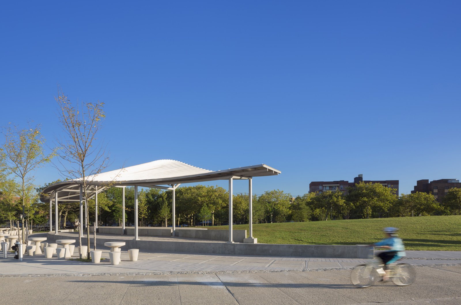 A minimalist pavilion with a wavy roof and open seating invites relaxation and recreation in an urban park.