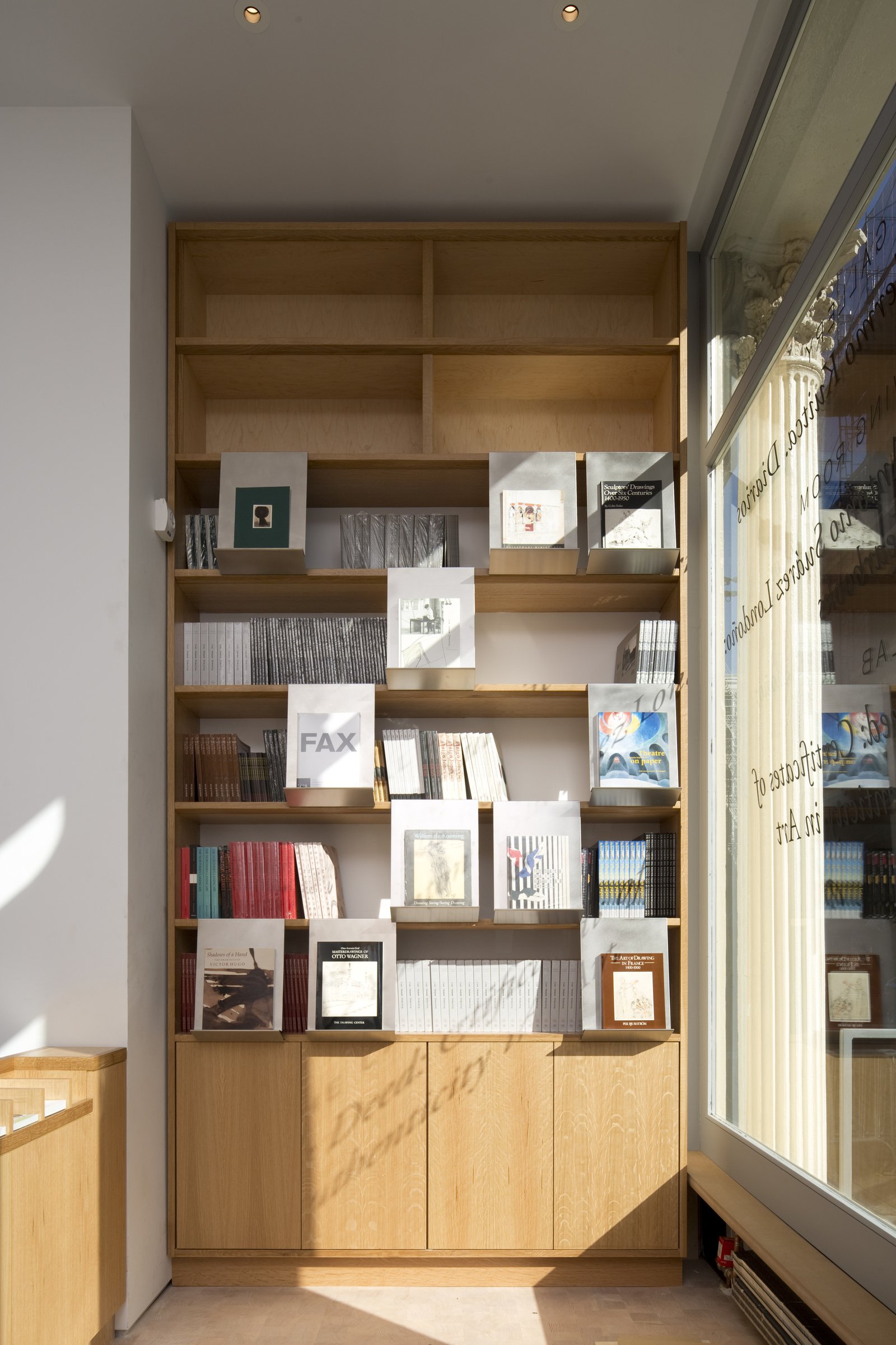 Built-in oak bookshelf displaying art books and catalogs, with afternoon light casting shadows through storefront window.