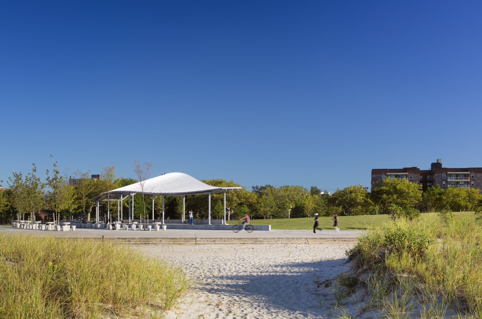 A performance canopy with a white roof and slender columns near sandy dunes and grassy parkland.