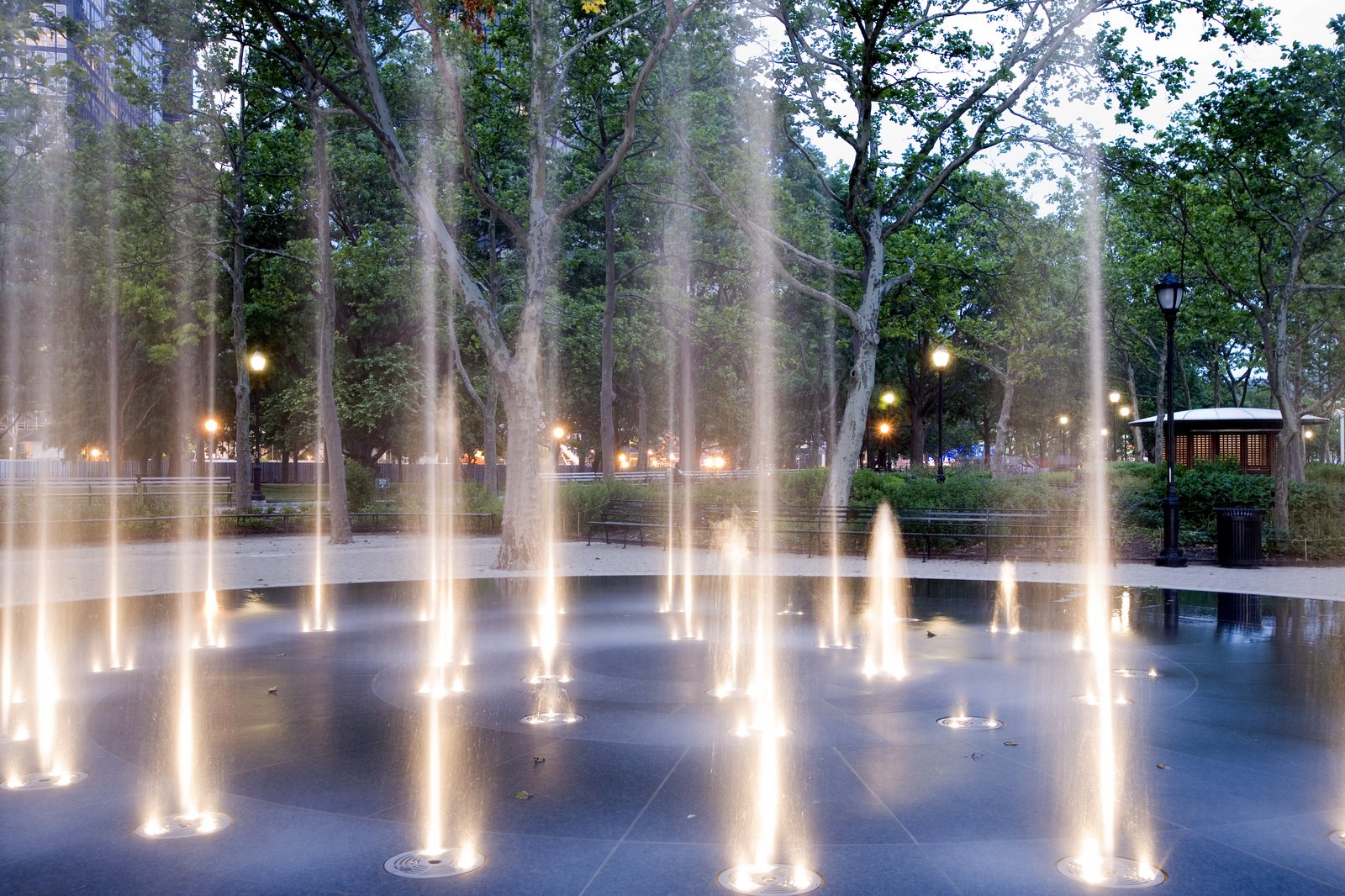 Illuminated fountain jets light up a park surrounded by trees at dusk.