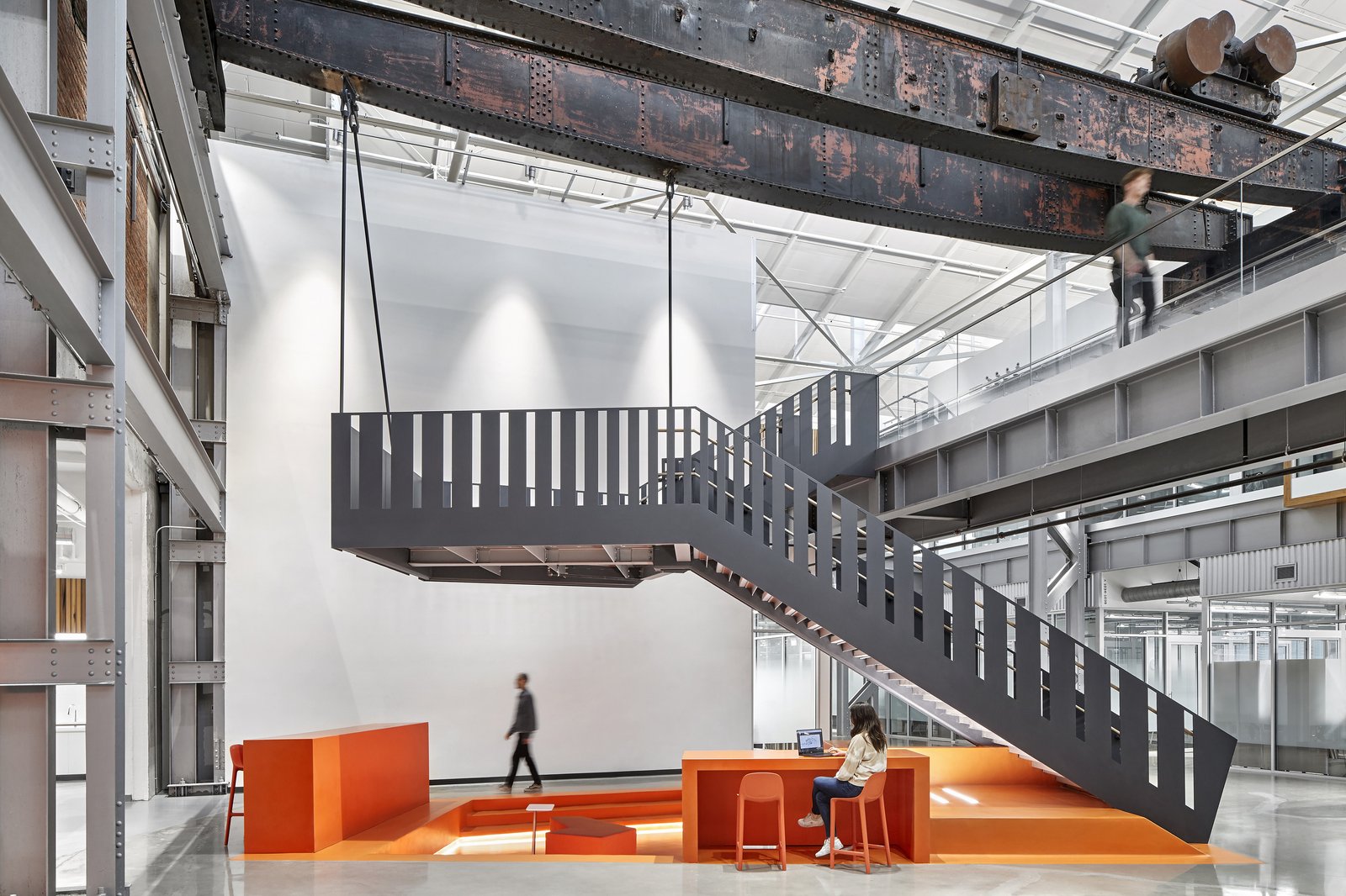 Industrial loft interior features a suspended steel staircase above orange seating area, with exposed beams.