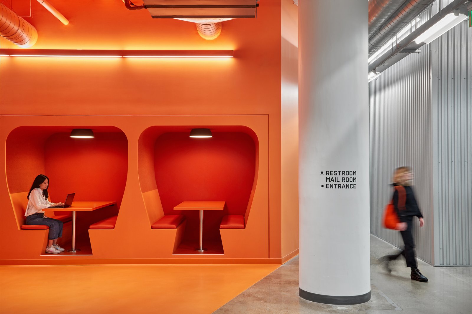 Modern office space with bright orange alcove seating booths and white column, showing wayfinding signage.