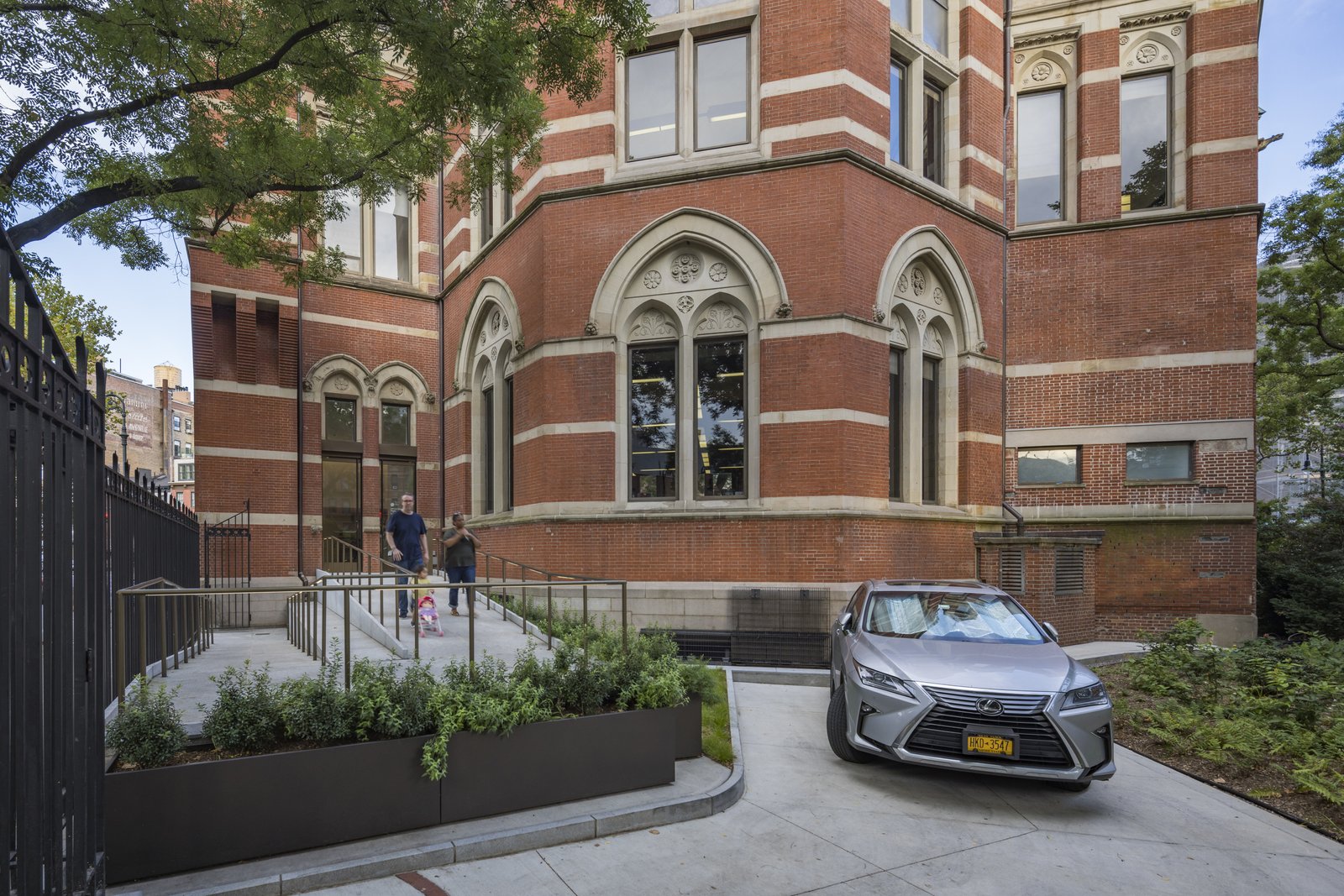 A curved driveway leads to a ramp entrance of a brick building with Gothic windows, bordered by raised garden beds.