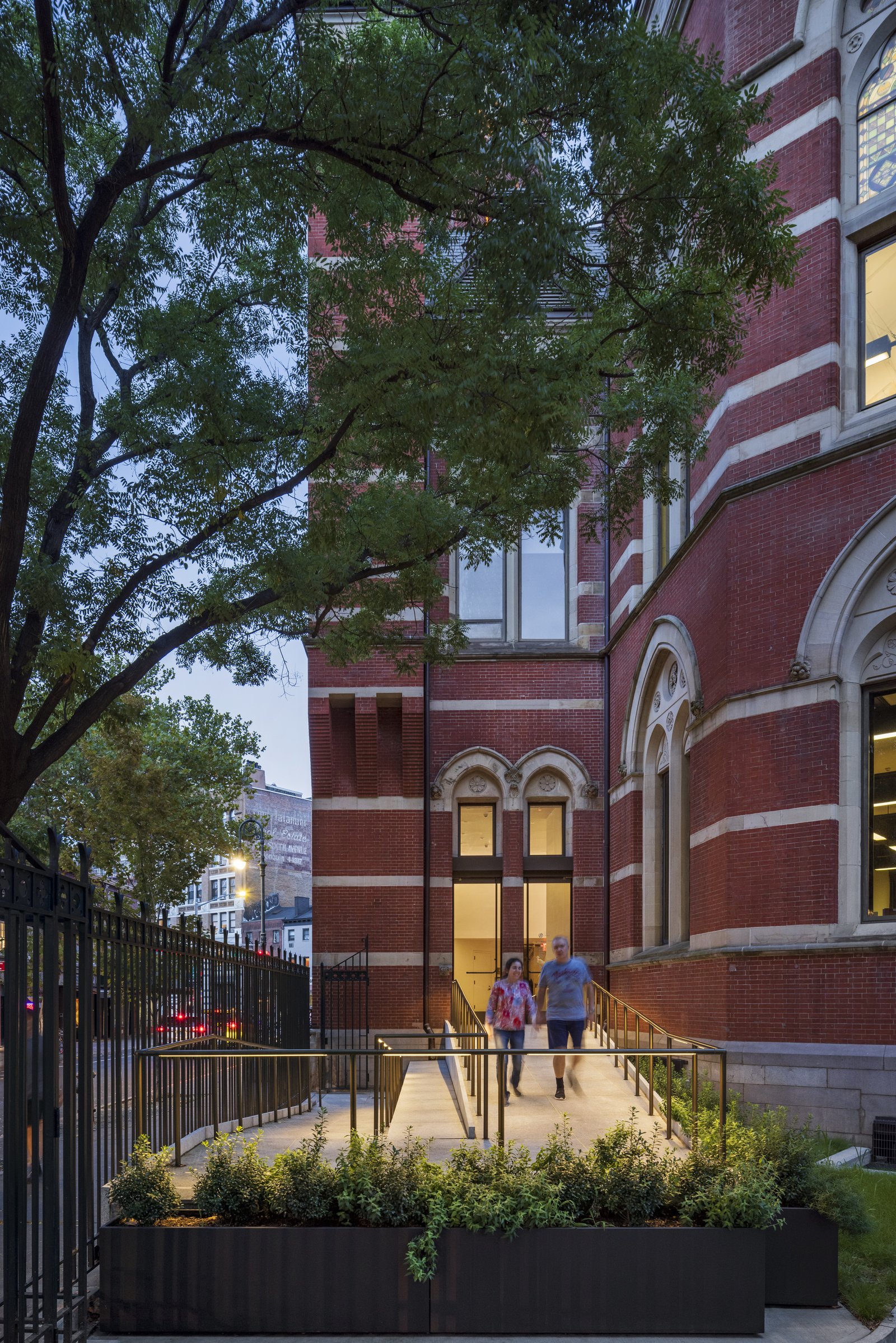 The exterior of the red brick library has a lit entrance with a ramp pathway surrounded by planters and trees at dusk.