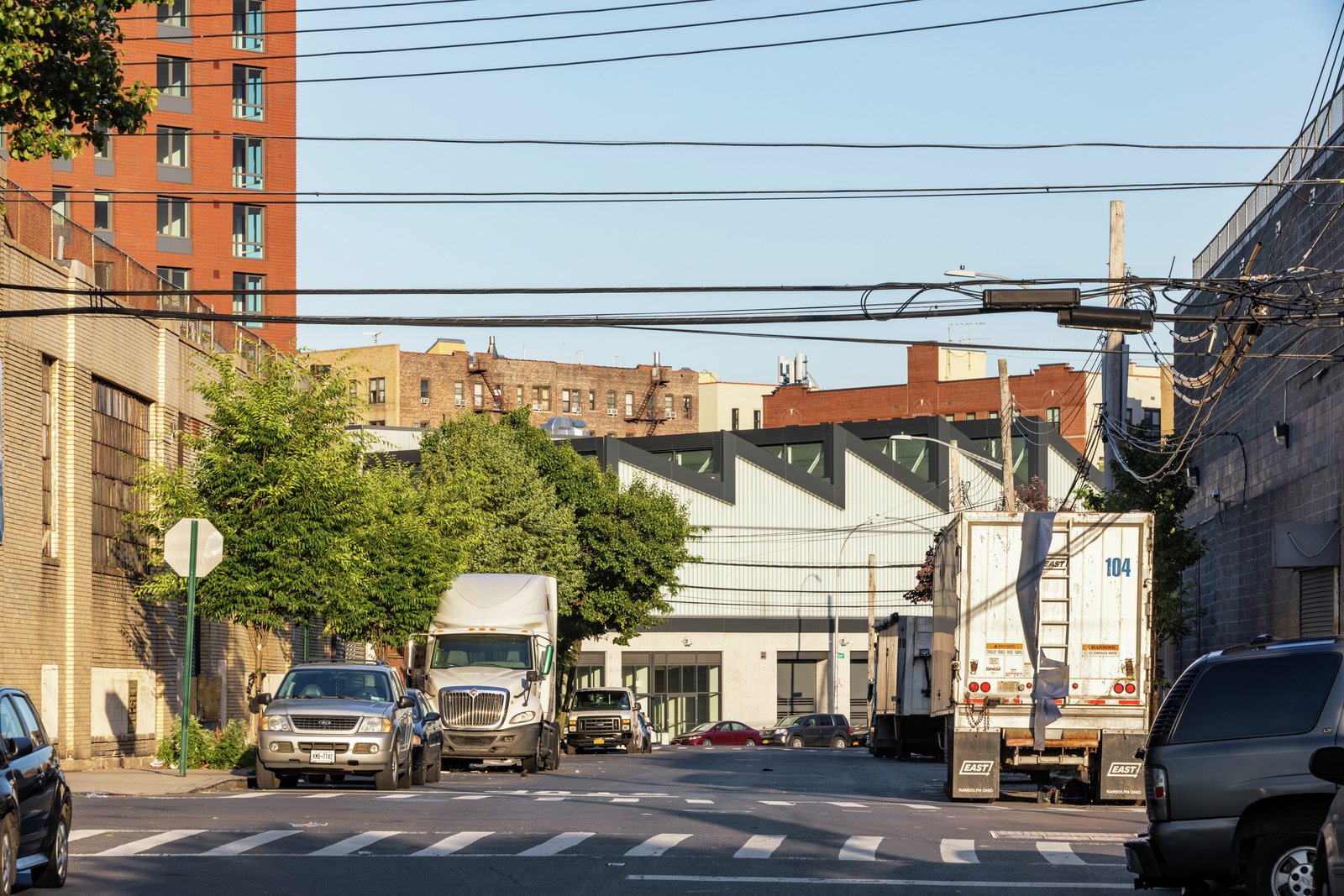 Street view of industrial building with sawtooth roof, framed by trucks and urban surroundings.