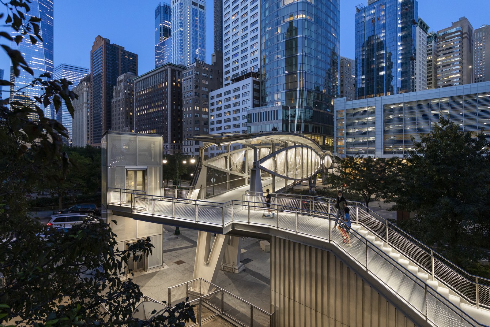 Elevated pedestrian bridge illuminated at dusk against the backdrop of city skyscrapers.