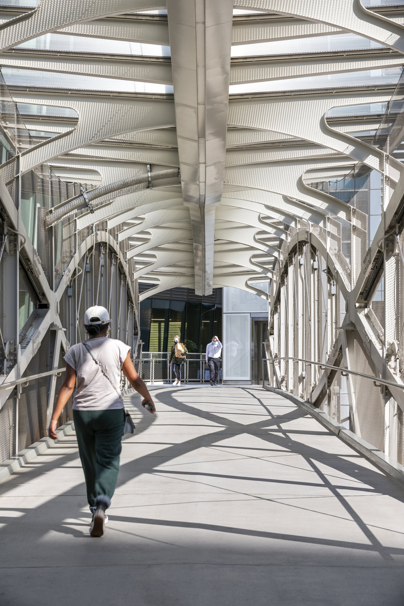 Person walking across a pedestrian bridge with open metal trusses and natural lighting.