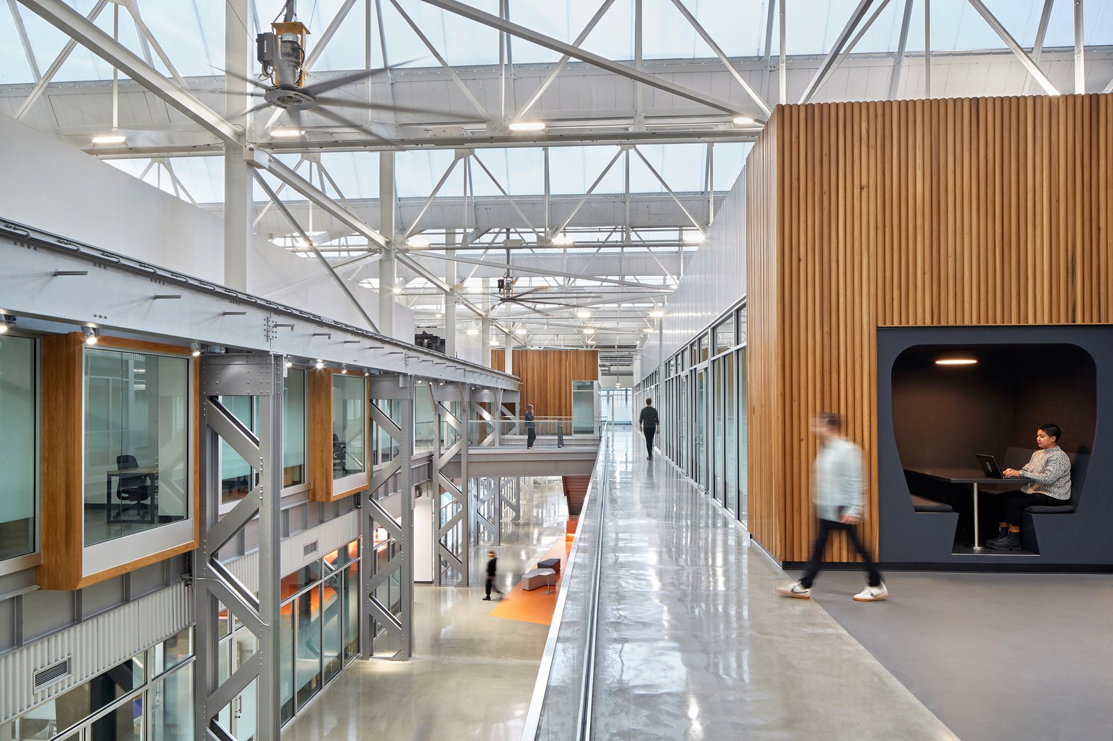 Double-height industrial workspace with exposed truss ceiling, wood-clad walls, and elevated walkway overlooking atrium.