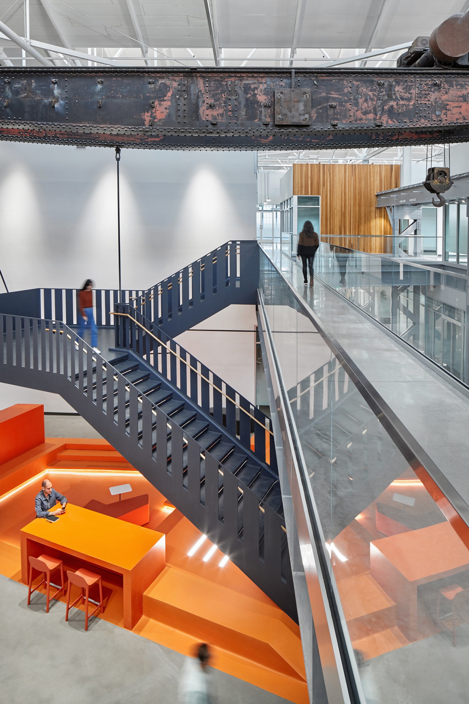 Overhead crane beam above modern staircase with glass railings, connecting orange seating area below."