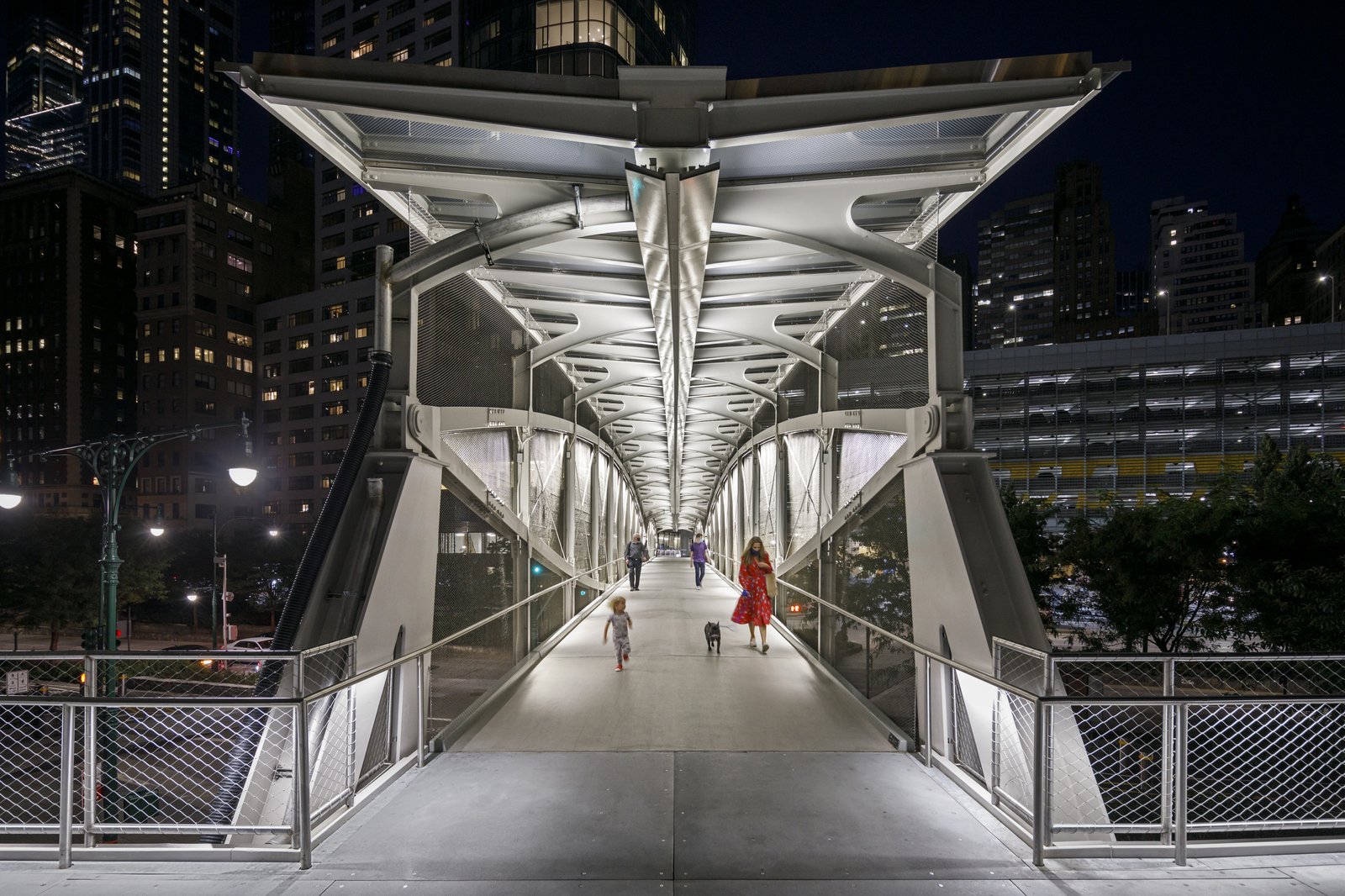 People walking across an illuminated pedestrian bridge at night with city lights in the background.