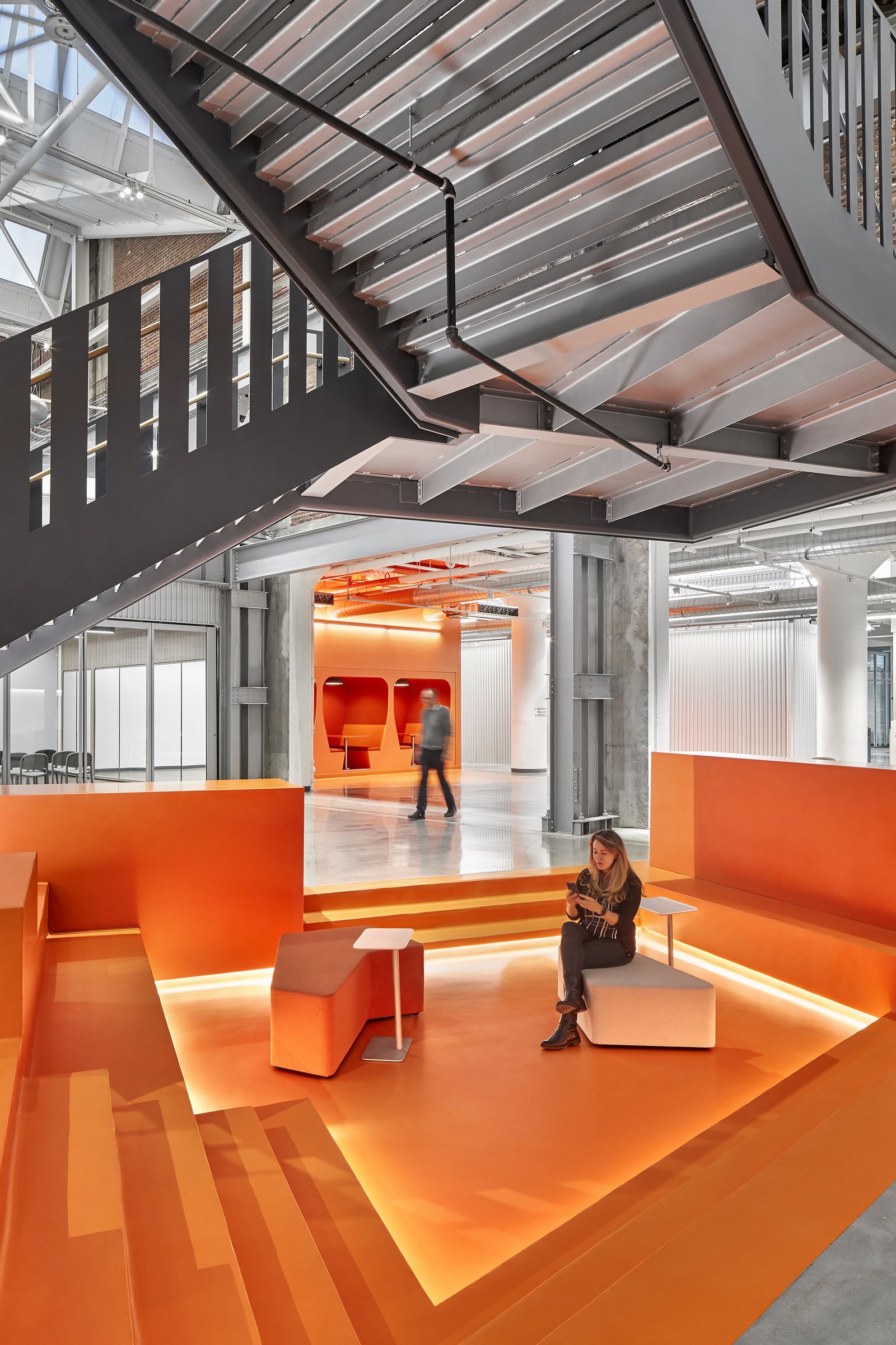 Tiered orange seating area beneath industrial staircase, with modern backlit steps and concrete columns in background.