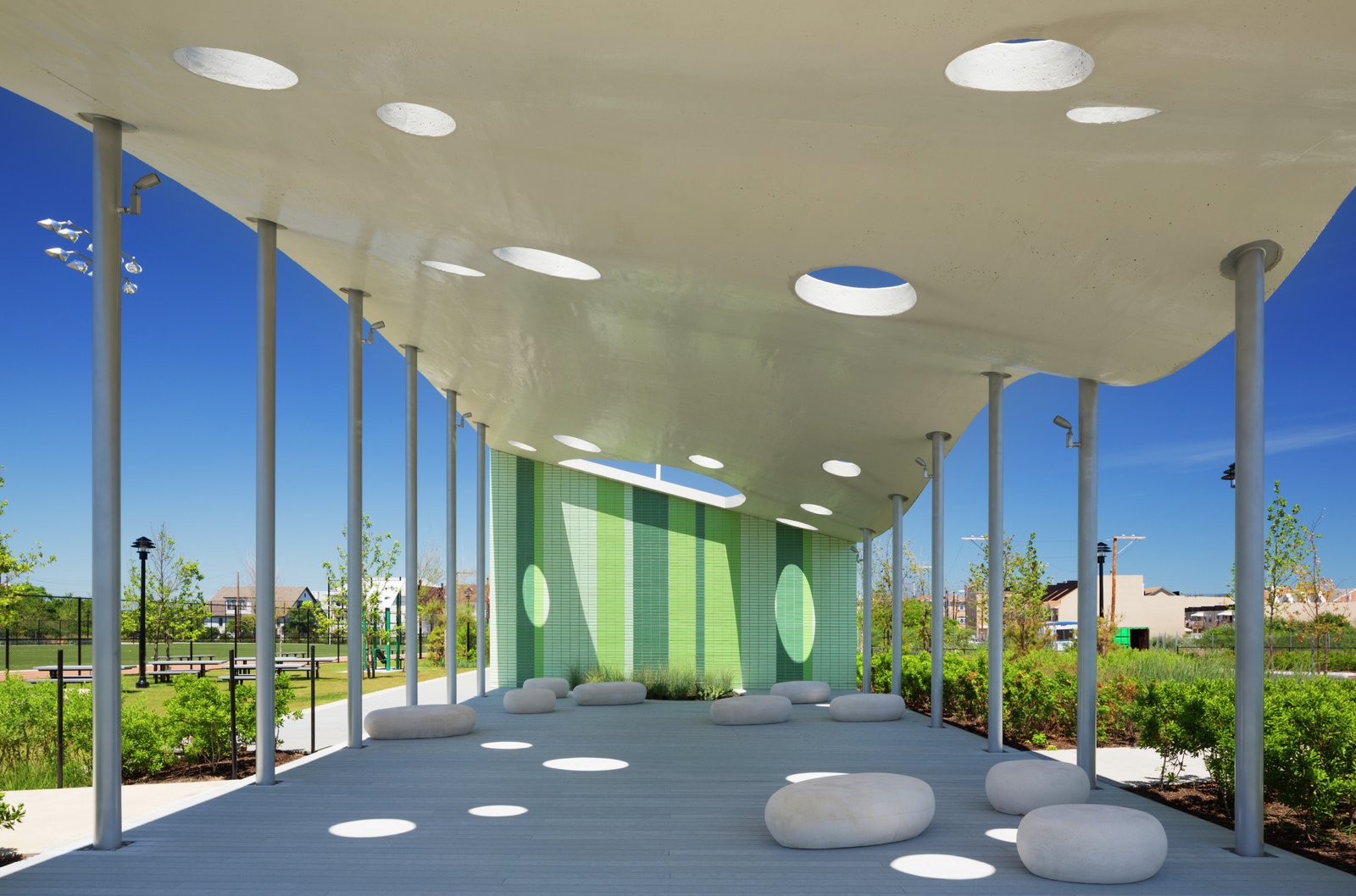 A pavilion with a perforated roof and green tiled wall offers shade and seating in a modern park setting.