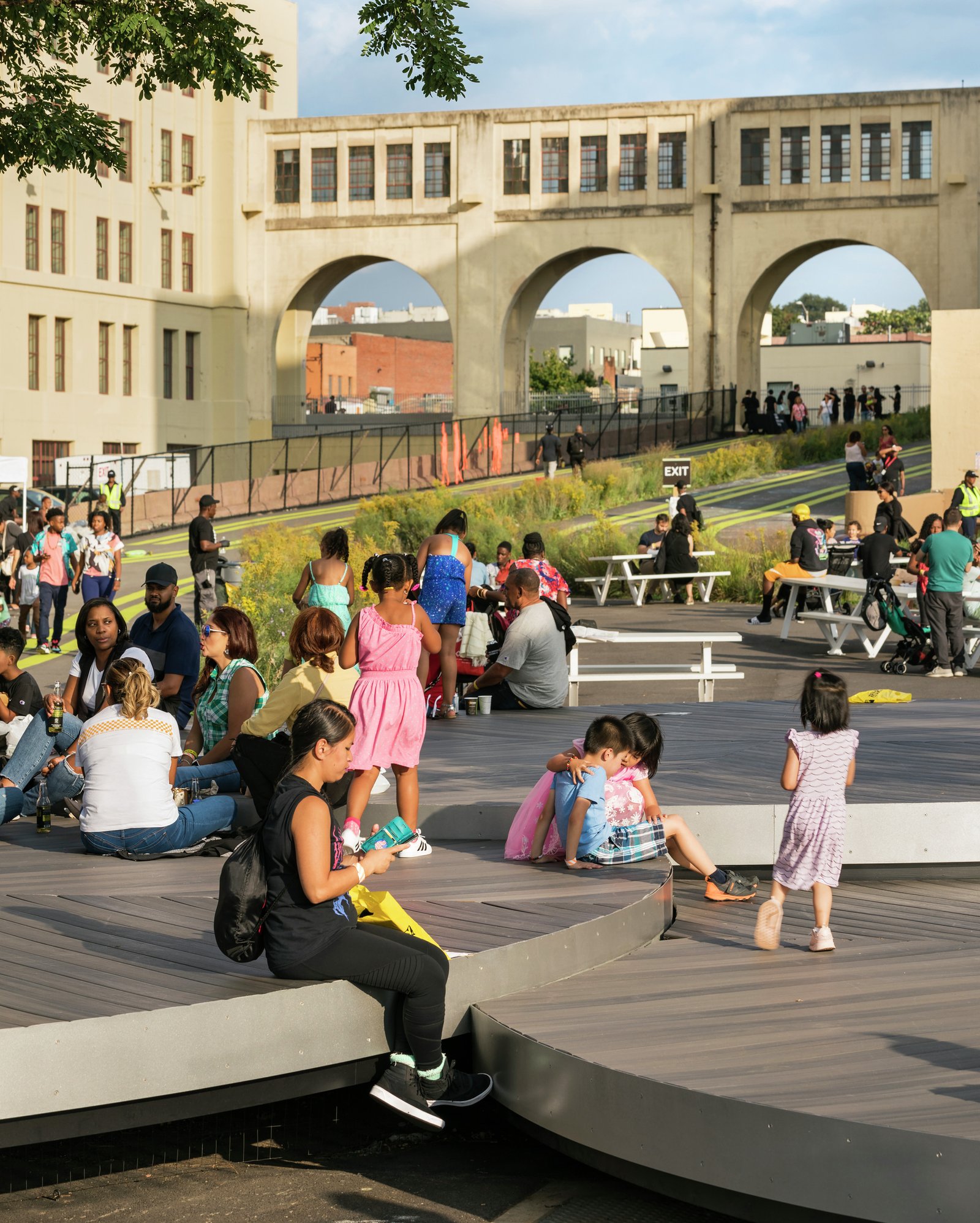 Families sit and children play on circular wooden seating near a historic arched bridge structure.