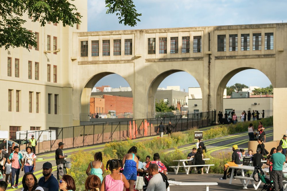 Families sit and children play on circular wooden seating near a historic arched bridge structure.