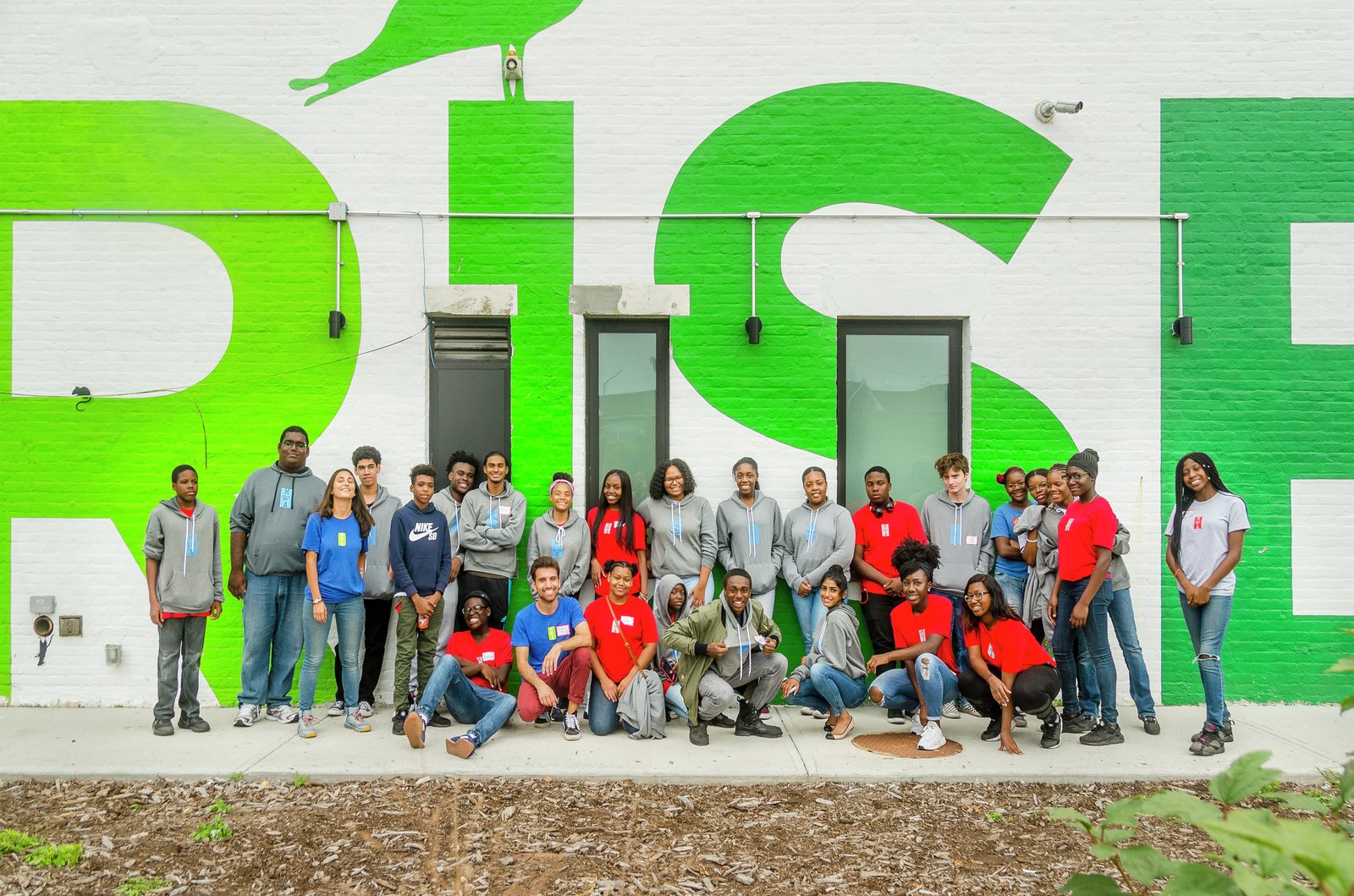 A diverse group of youth and community members pose in front of a building with a large "RISE" mural.