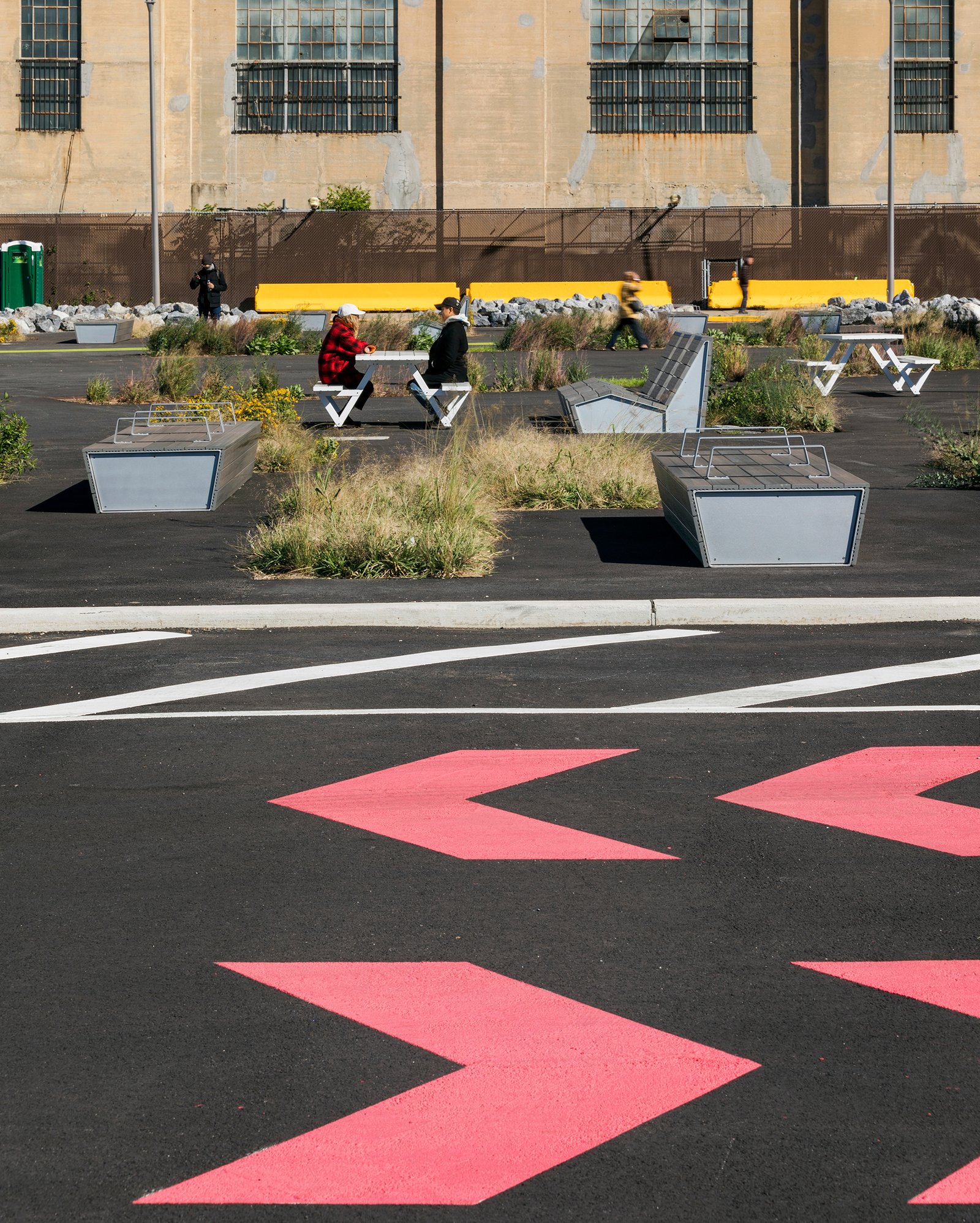 Bright pink arrows on asphalt lead to seating areas surrounded by greenery and an industrial facade.