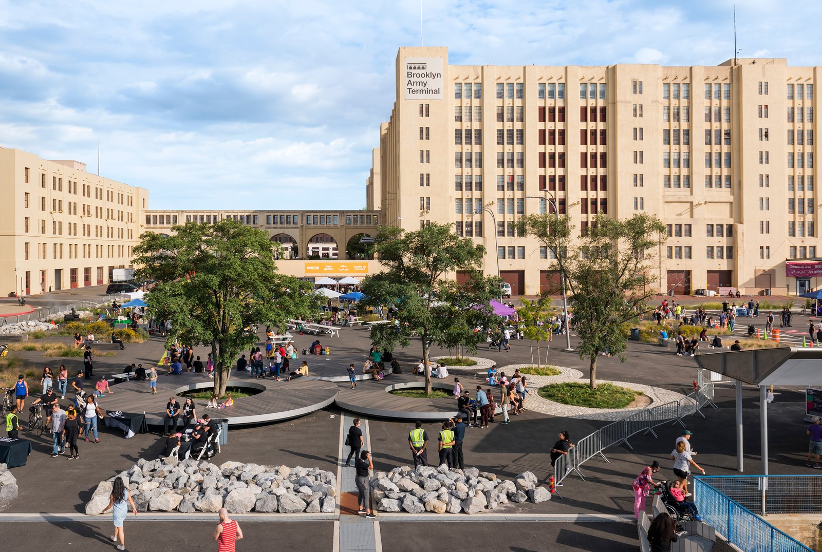 A bustling public plaza with trees, circular wooden seating, and people enjoying the open space.