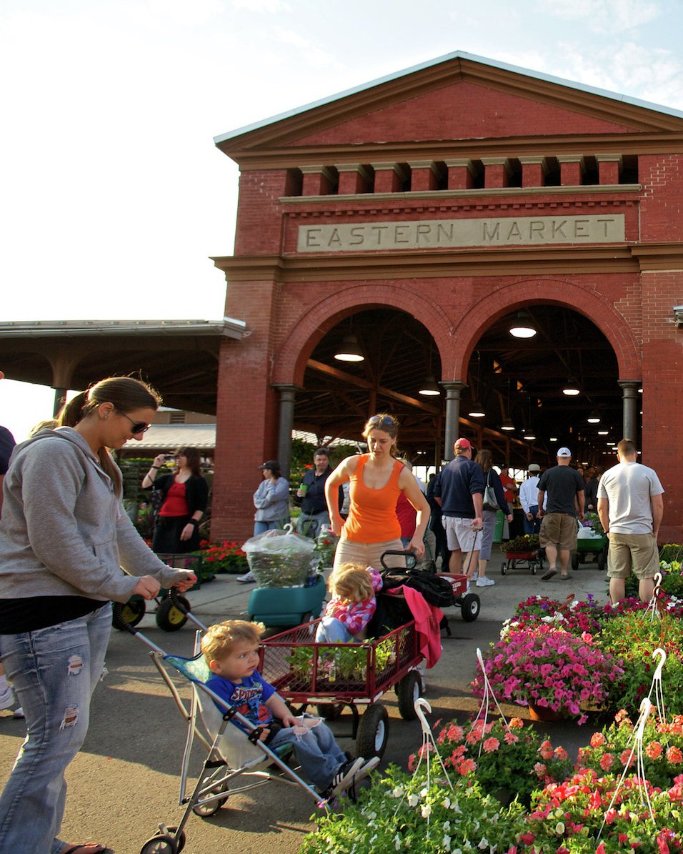 A saxophone player performs in front of the bustling Eastern Market with flowers and families nearby.