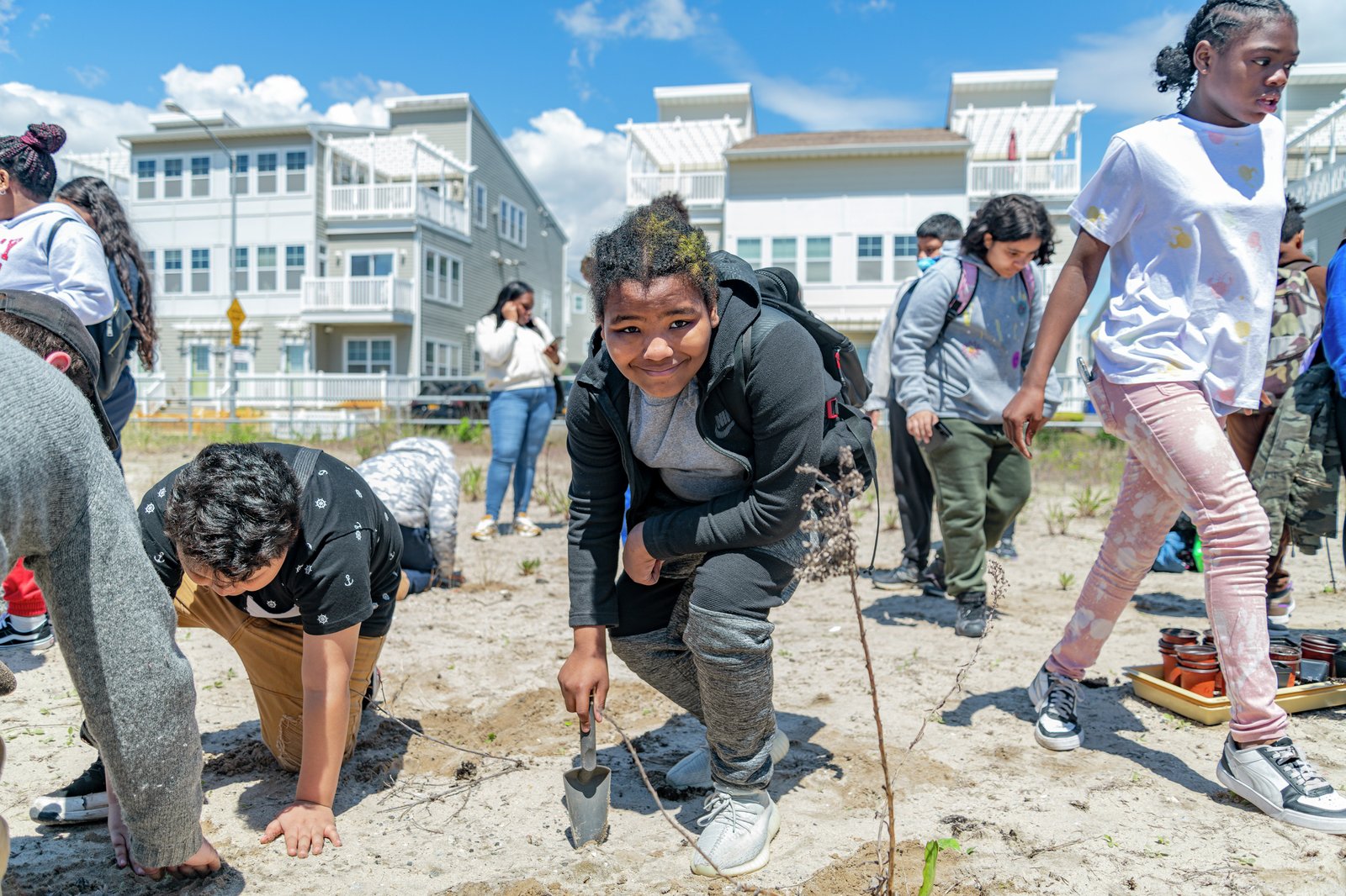 Children planting grasses in sandy soil with houses and clear blue skies in the background.