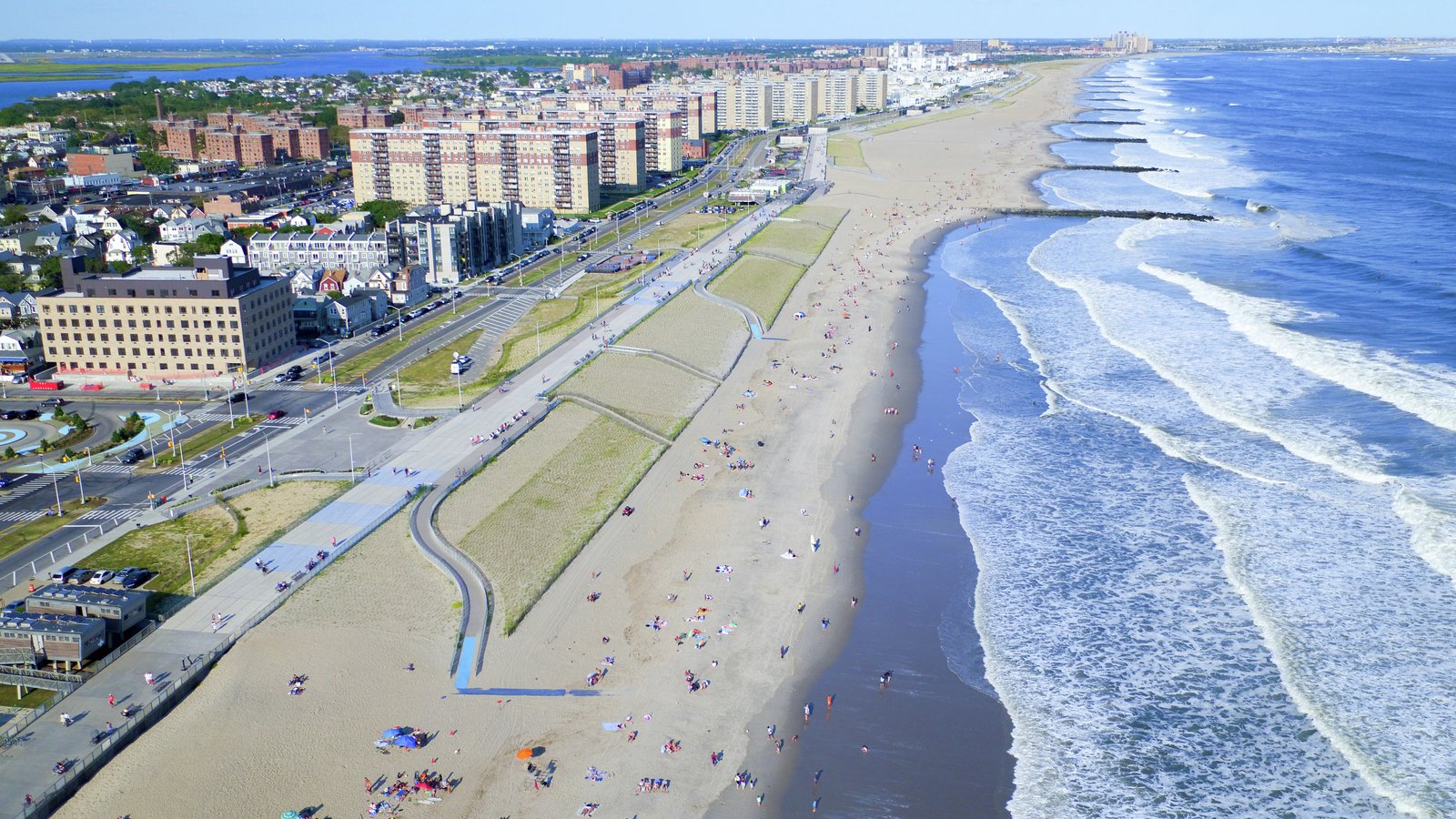 Aerial view of a beach, boardwalk, and urban backdrop, with pathways, dunes, and people enjoying the vibrant shoreline.