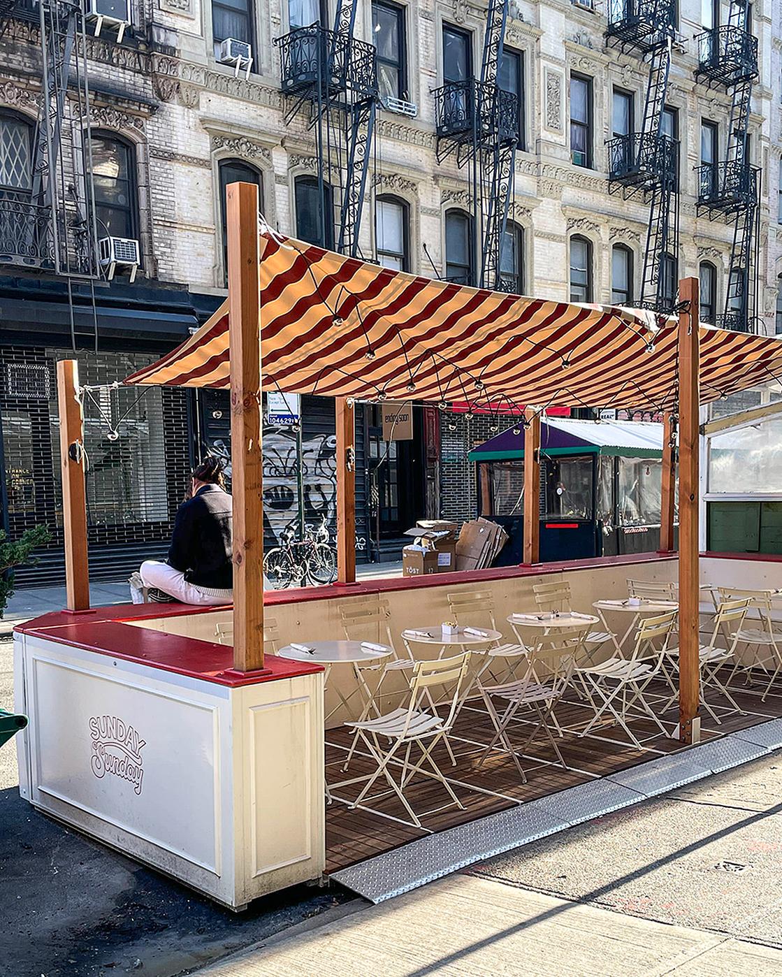 Street-level outdoor dining with striped canopy, tables, and chairs on a busy urban street.