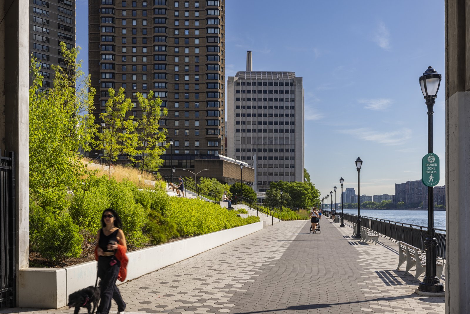 A riverside walkway with benches, greenery, and city buildings in the background.