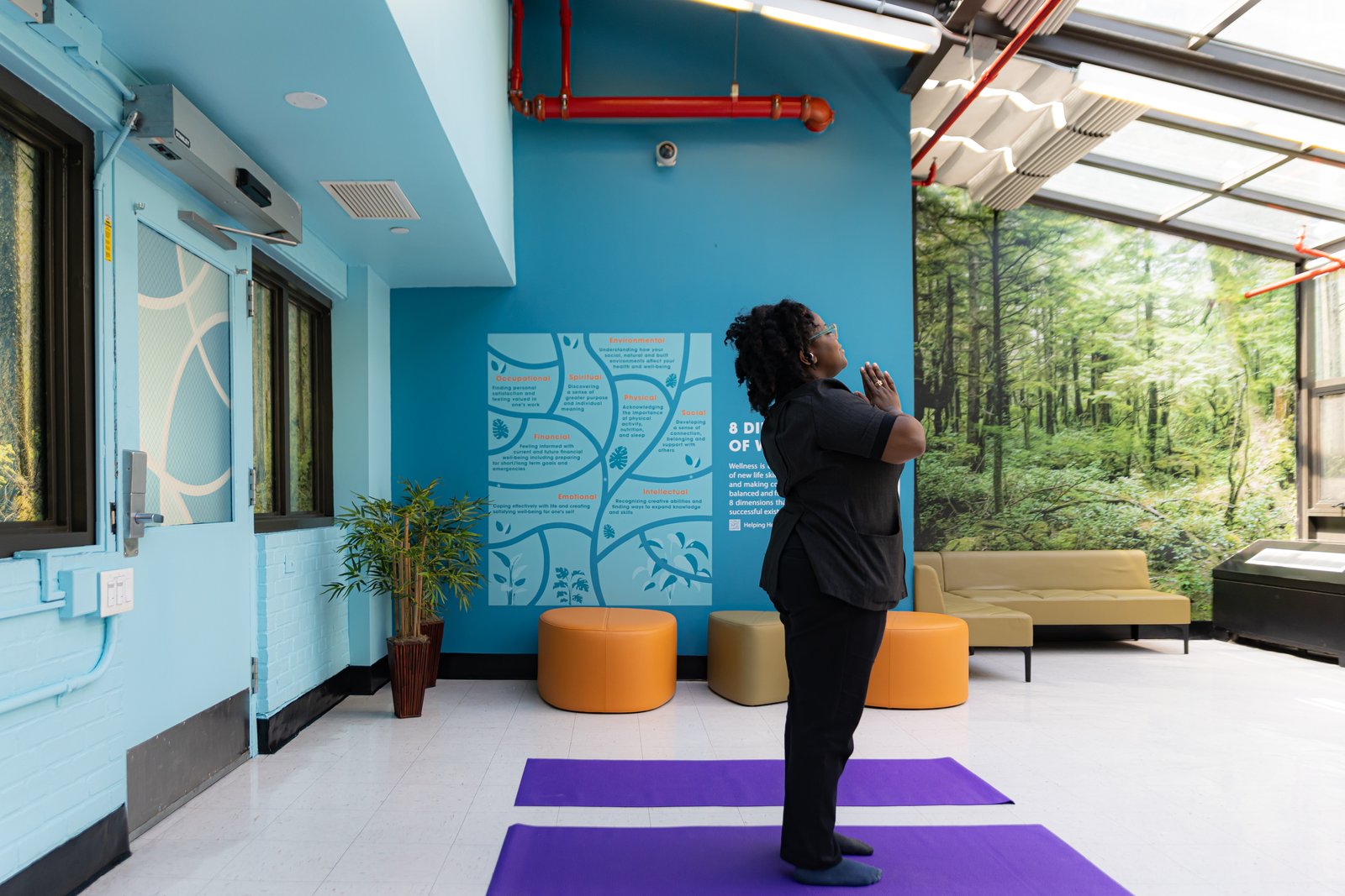 A wellness room with a person standing on a yoga mat, surrounded by forest murals, soft blue walls, and natural light.