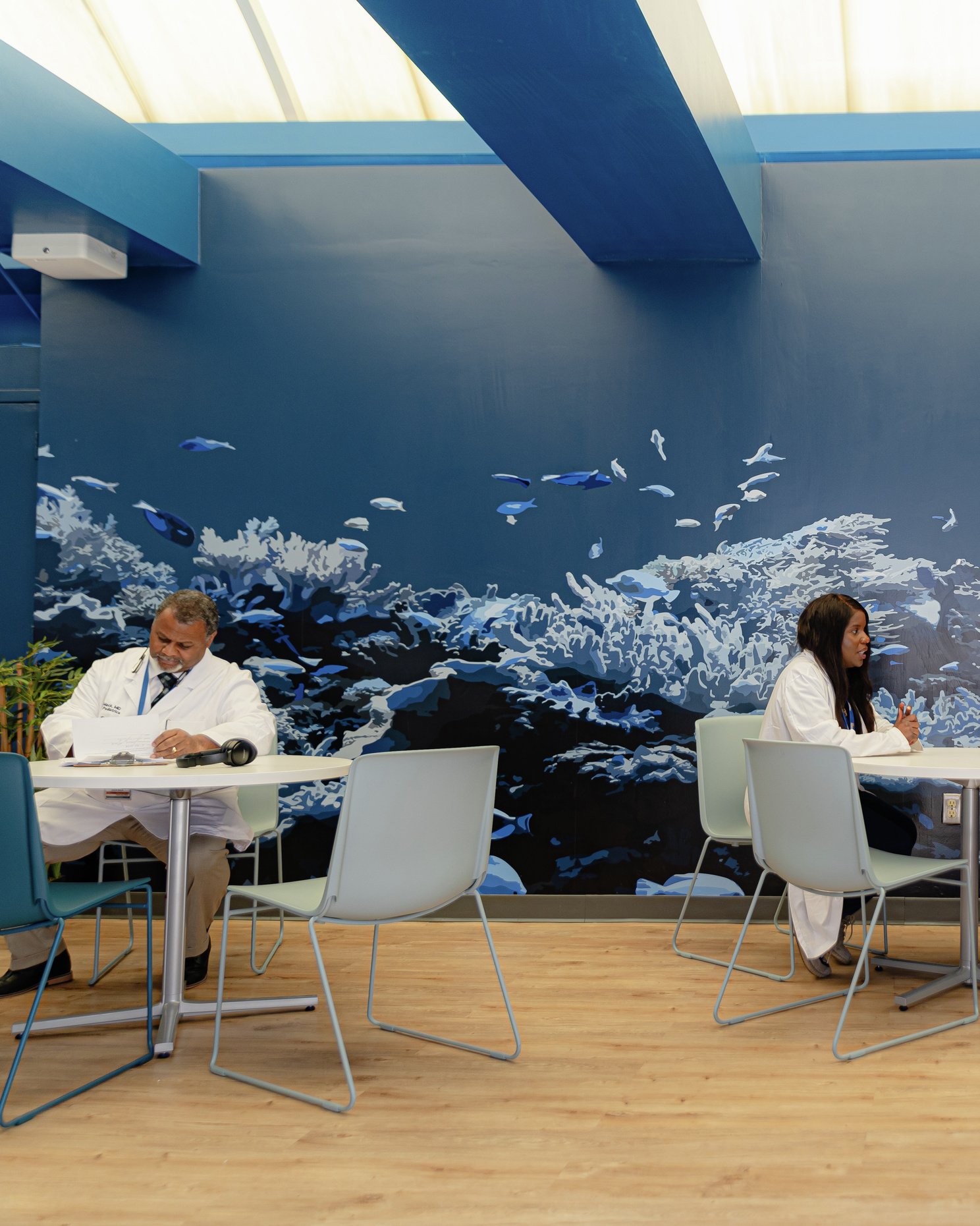 A wellness room featuring staff seated at tables, a vibrant ocean-themed mural, and soft blue beams with abundant natural light.