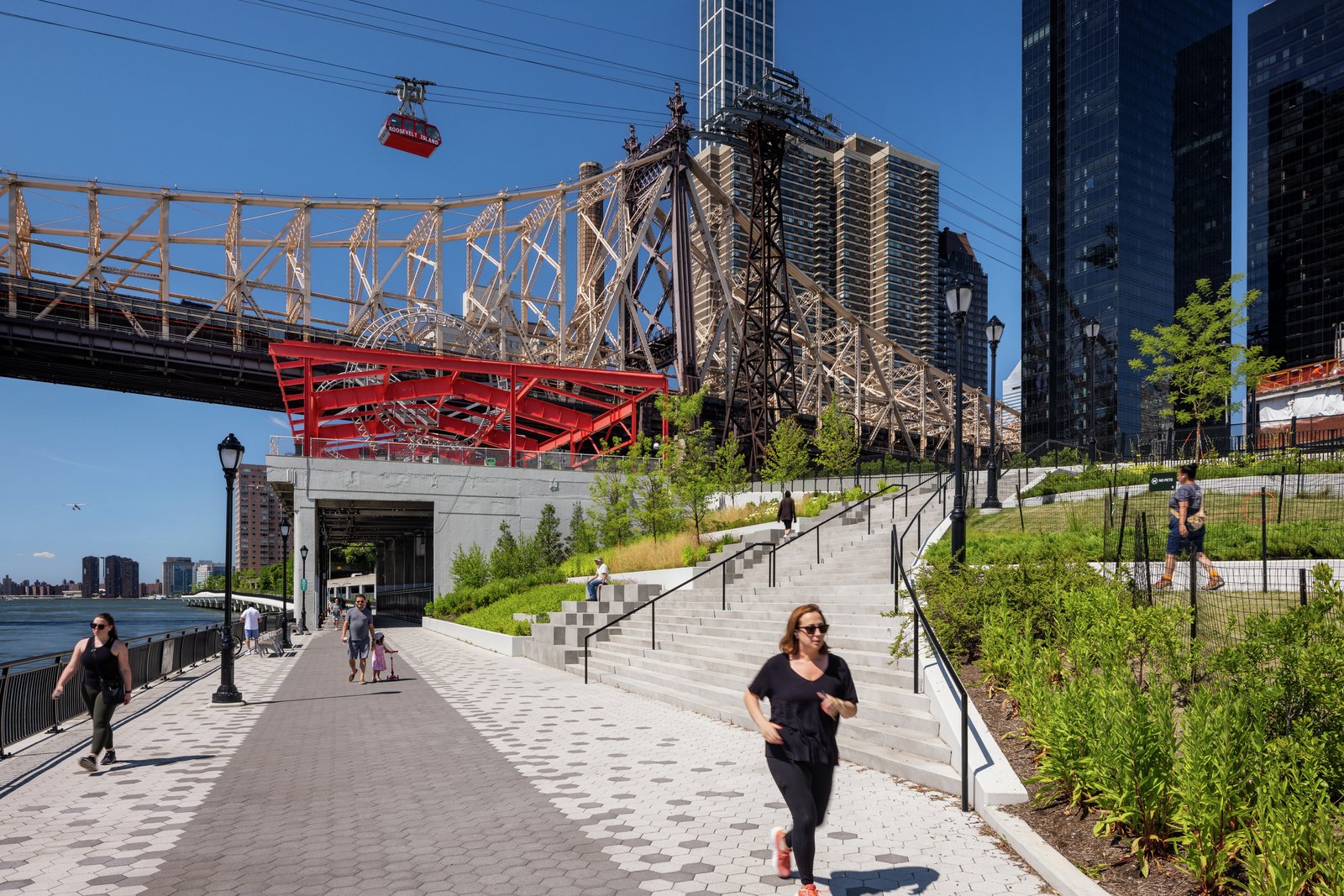 Wide riverside walkway with hexagonal paving, stairs, and a bridge overhead.