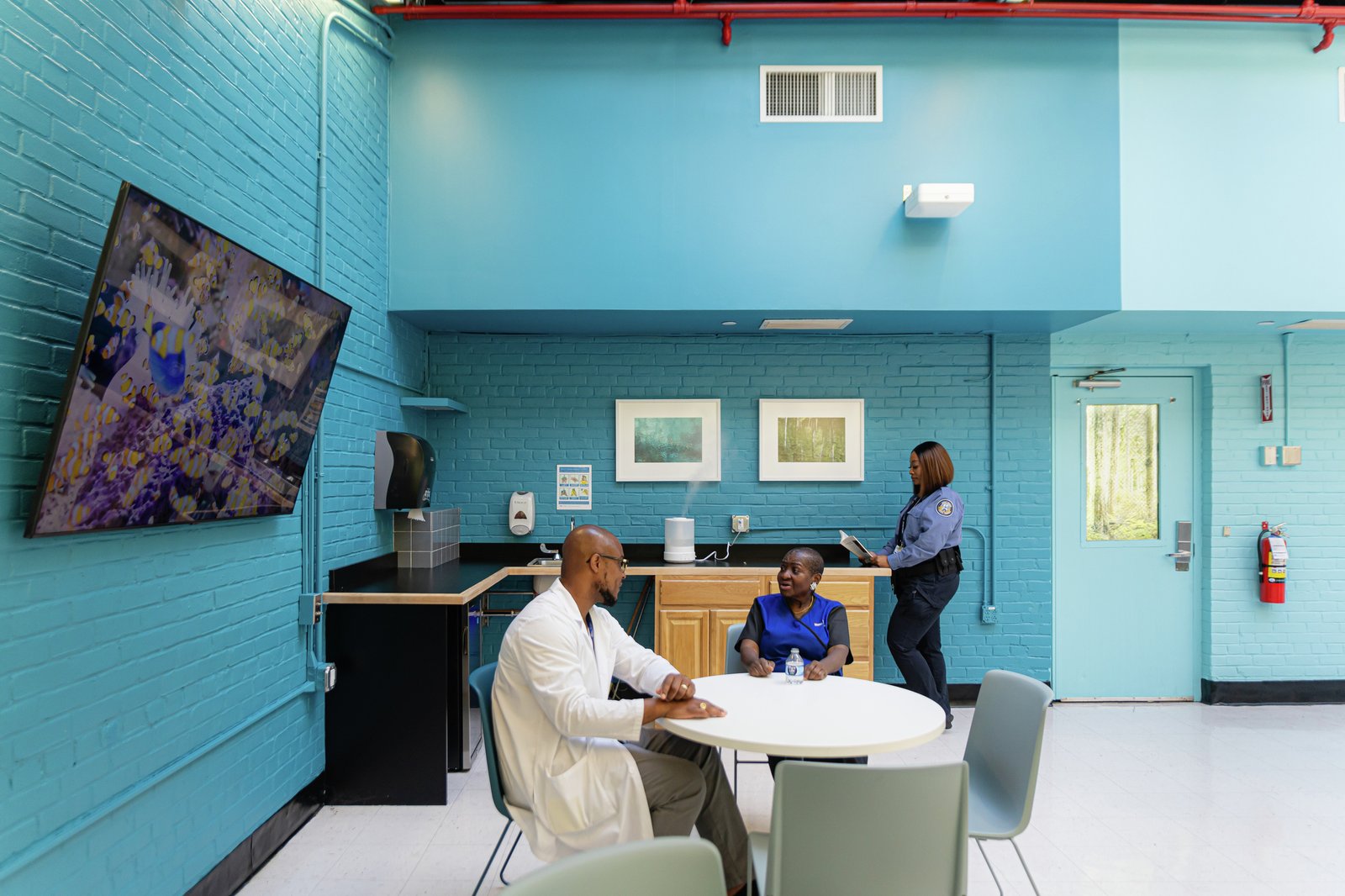 A wellness room with staff seated at a round table, surrounded by calming blue walls, framed artwork, and a kitchenette area.