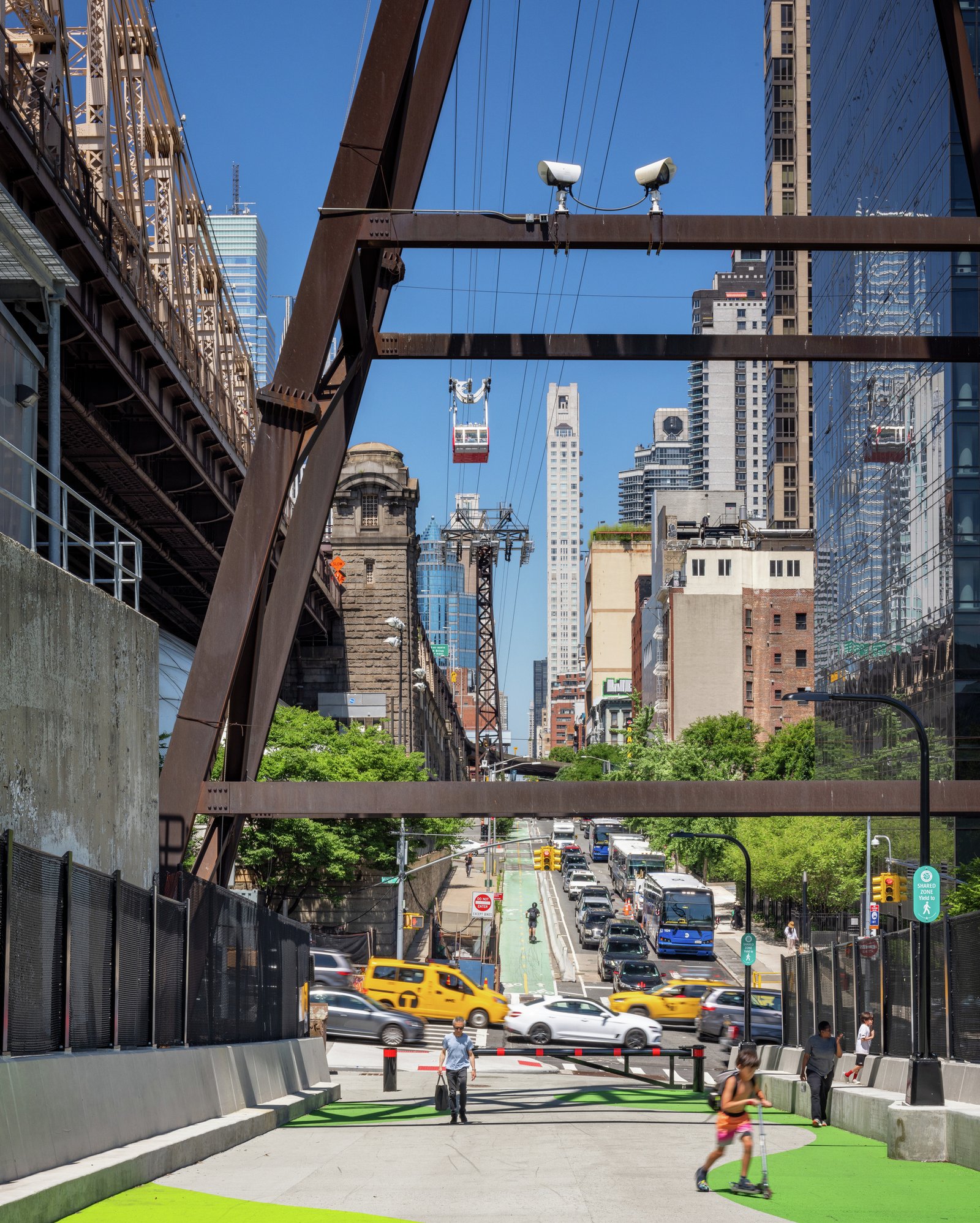 Pathway under steel supports with Roosevelt Island tram overhead and urban skyline visible.